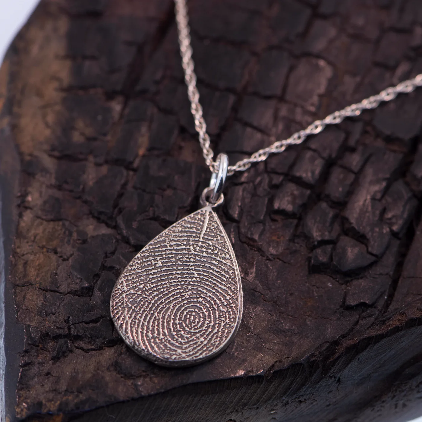 A silver teardrop pendant carrying a full fingerprint, on charred wood